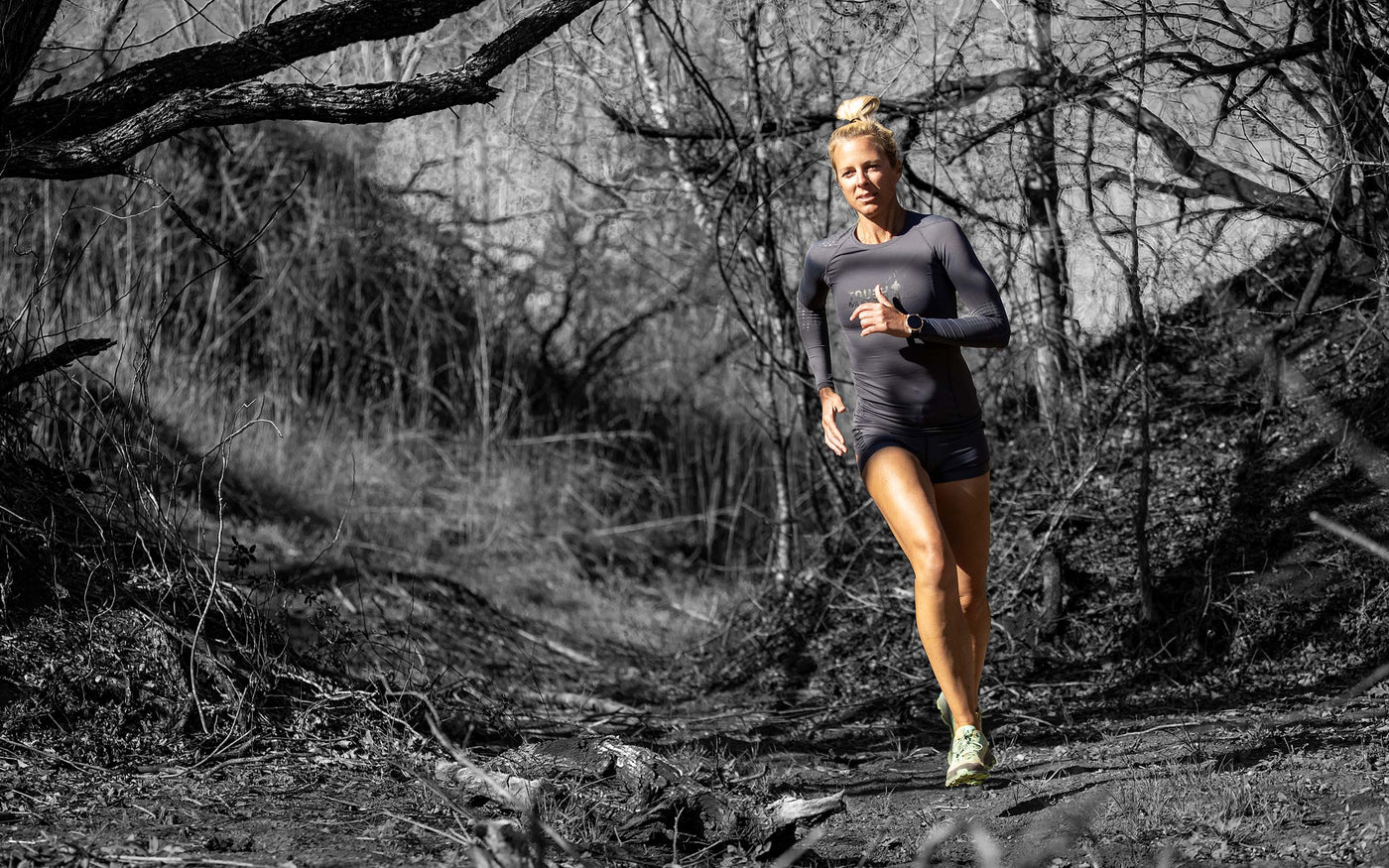 Tough Mudder female athlete trail running through the woods wearing a craft performance long sleeve top and shorts